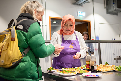 A female entrepreneur serving food to a member of the public at an indoor food stall.